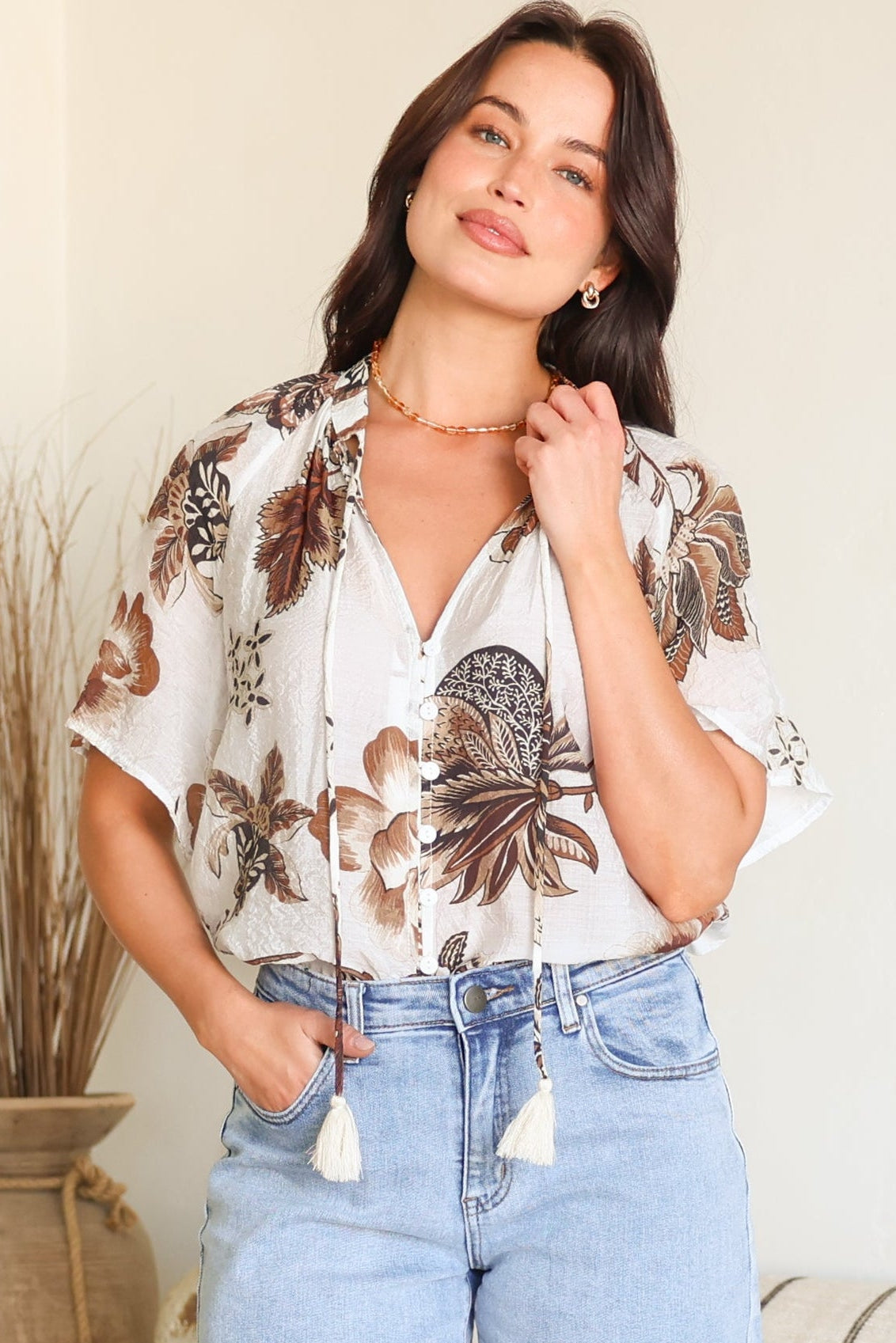 A woman modeling a white short-sleeve blouse with a brown and beige botanical print, paired with light-wash denim shorts, standing in a brightly lit indoor setting.