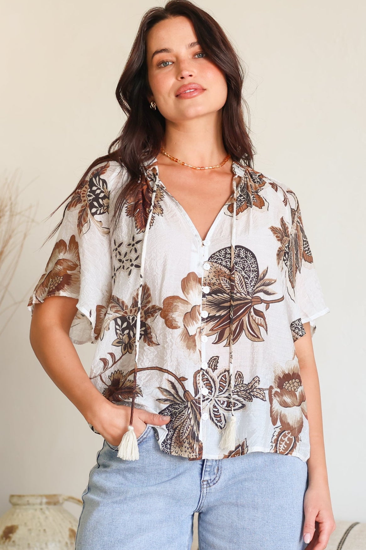 A woman wearing a white short-sleeve button-up blouse with a brown and beige floral print, paired with light-wash denim shorts, standing in a brightly lit indoor setting.