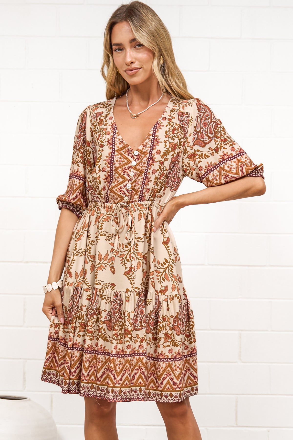 A woman in a tan paisley print mini dress with balloon sleeves, a V-neckline, tie-waist detail, and tiered skirt, posed against a white painted brick wall.