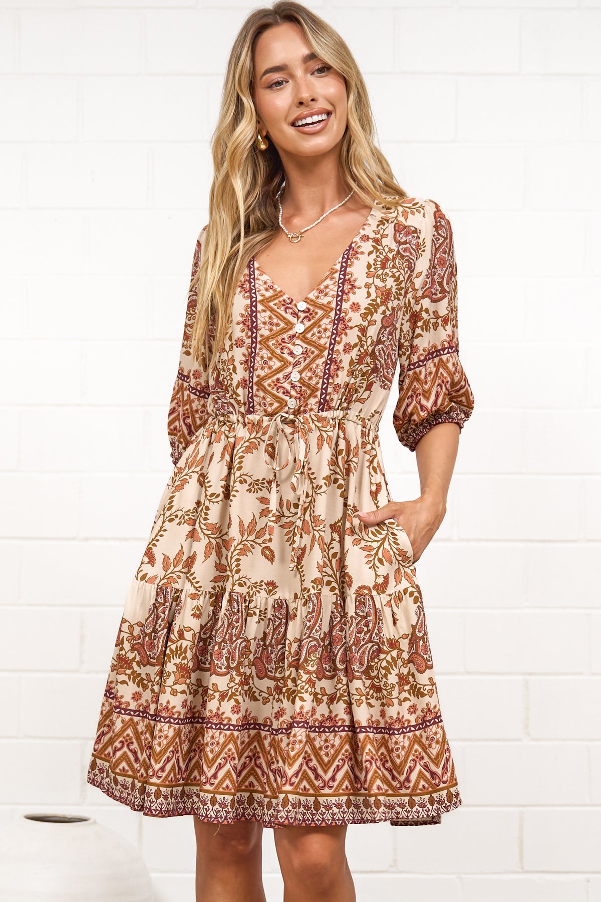 A woman in a tan paisley print mini dress with balloon sleeves, a V-neckline, tie-waist detail, and tiered skirt, posed against a white painted brick wall.