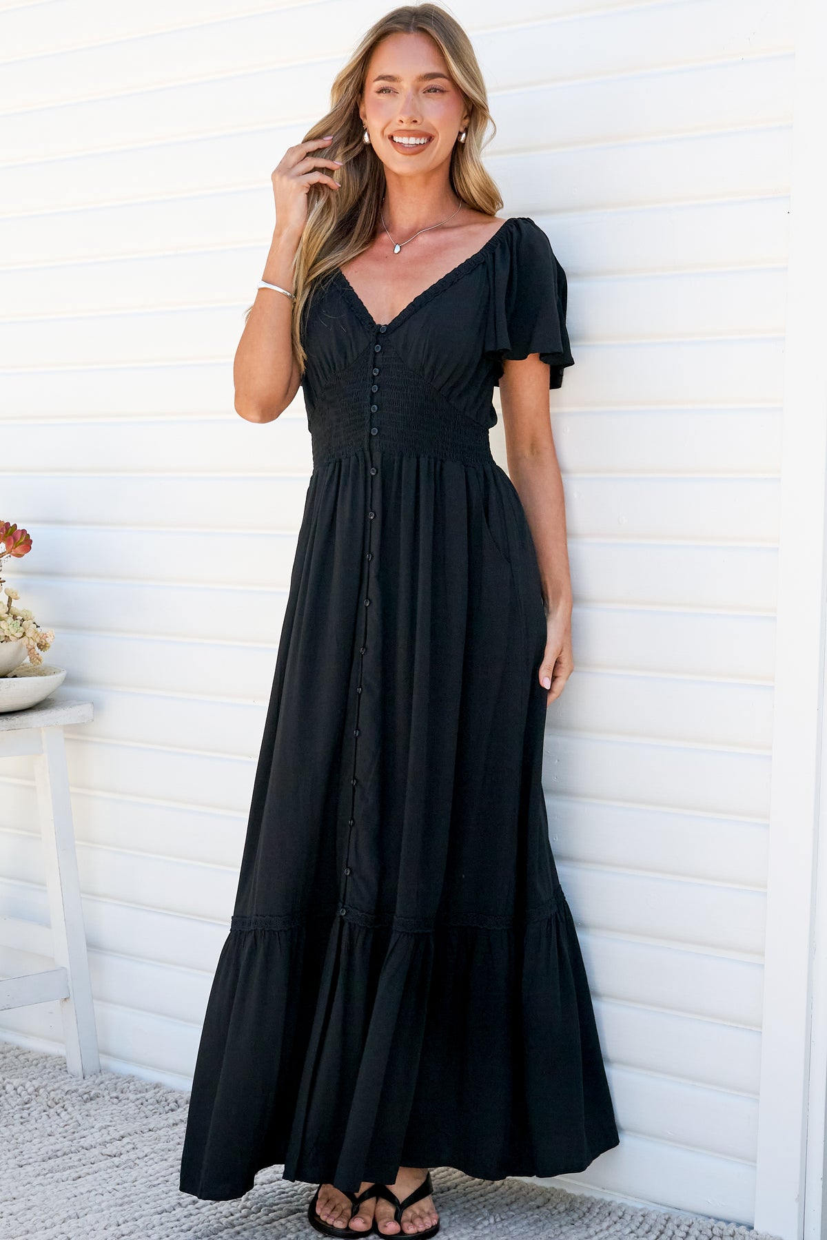 A smiling woman with one hand in her hair leaning against a white wall in the Marilyn Maxi Dress in Black, a floor-length dress with a smocked button-front bodice, flutter sleeves and lace-trimmed tiered hem