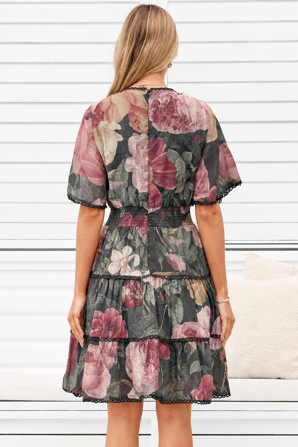 A woman viewed from the back wearing a tiered floral dress with short flutter sleeves and black lace trim, standing in front of a white paneled wall.