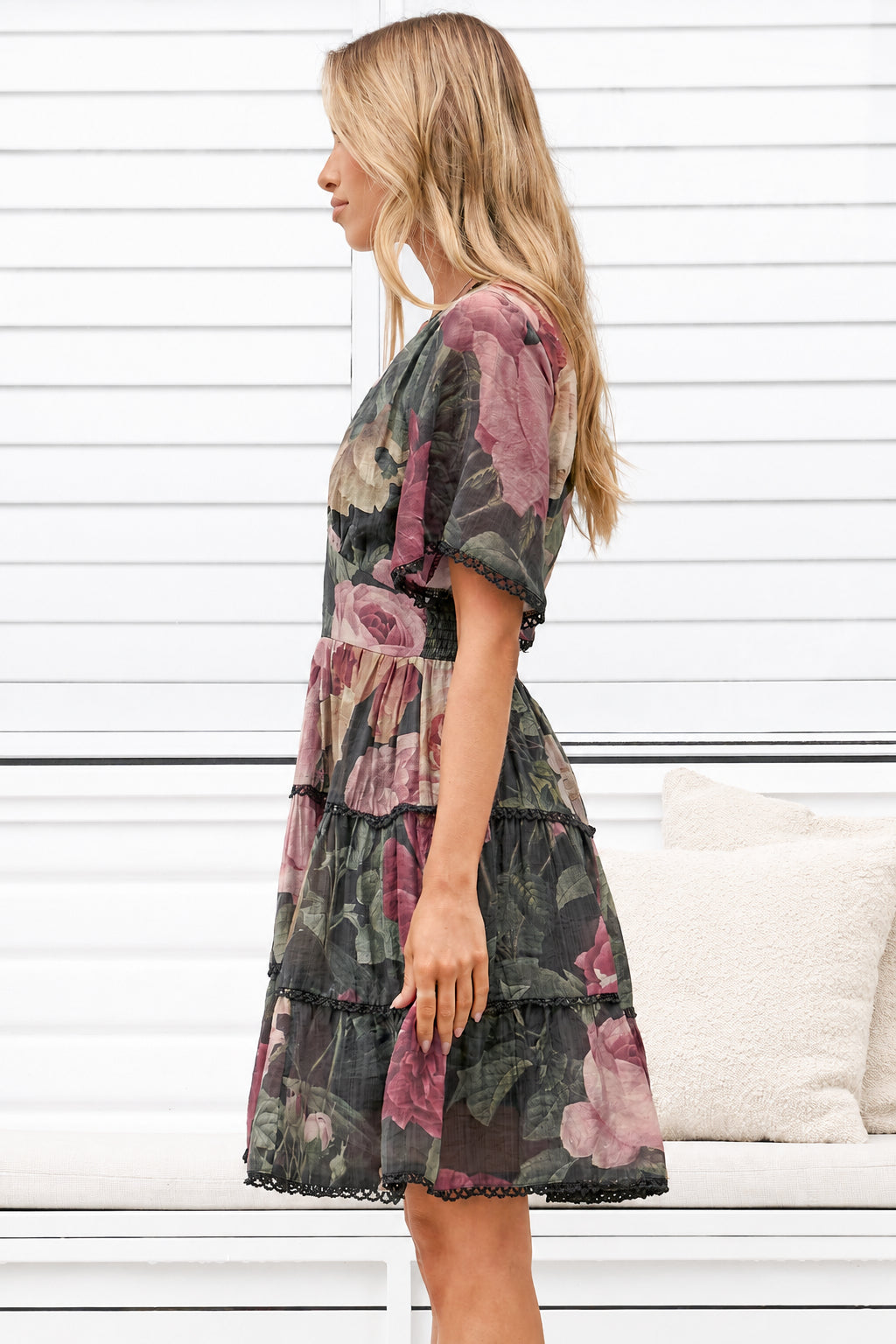 A woman wearing a short puff-sleeve tiered dress with a dark floral print, shown in profile against a white paneled wall.