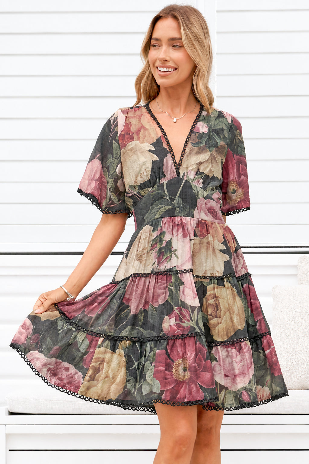 A woman wearing a tiered short-sleeve dress with a dark floral print, standing in front of a white paneled wall.