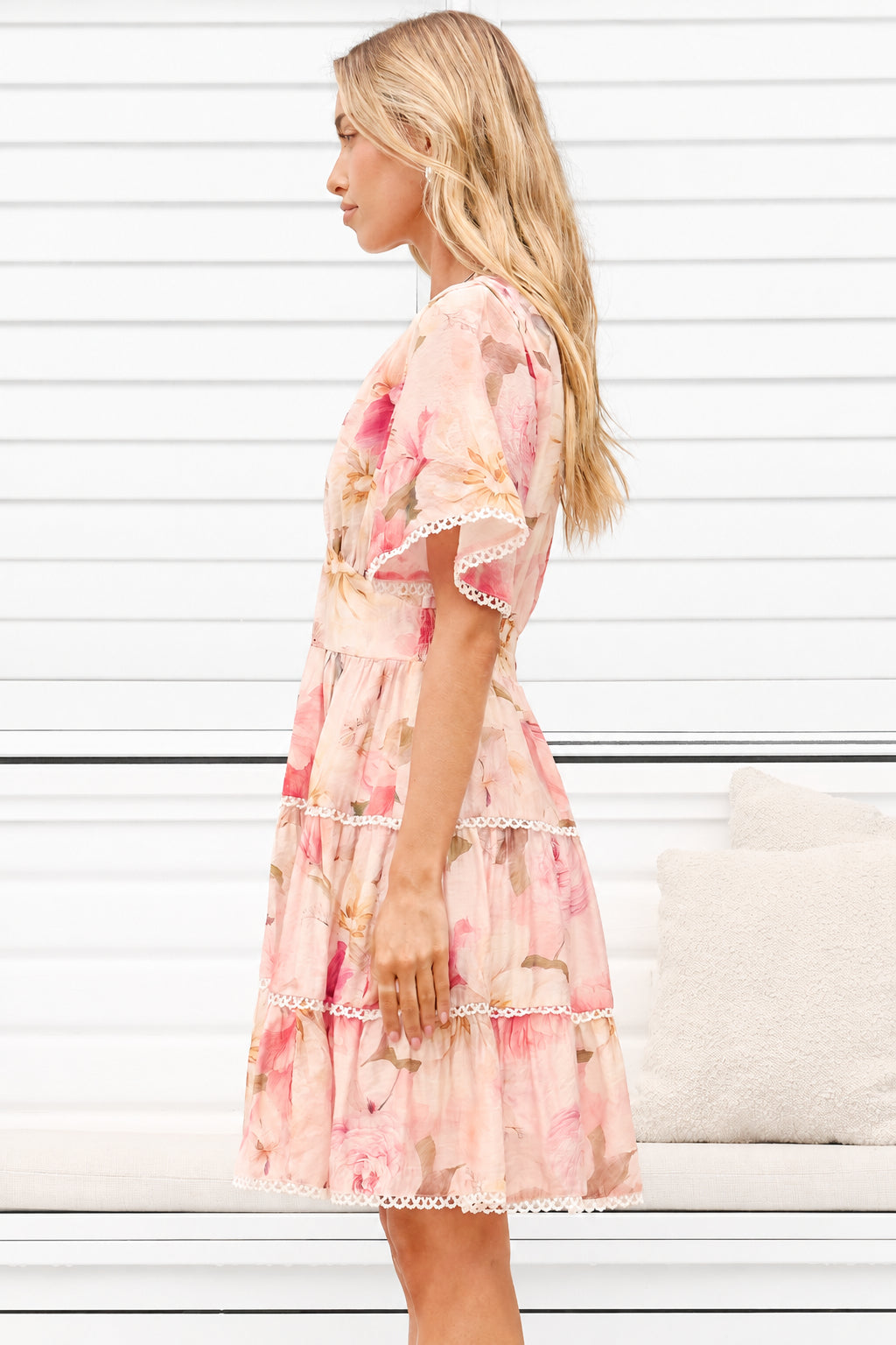 A woman wearing a light pink tiered dress with a floral print and white lace trim, shown in profile against a white paneled wall.