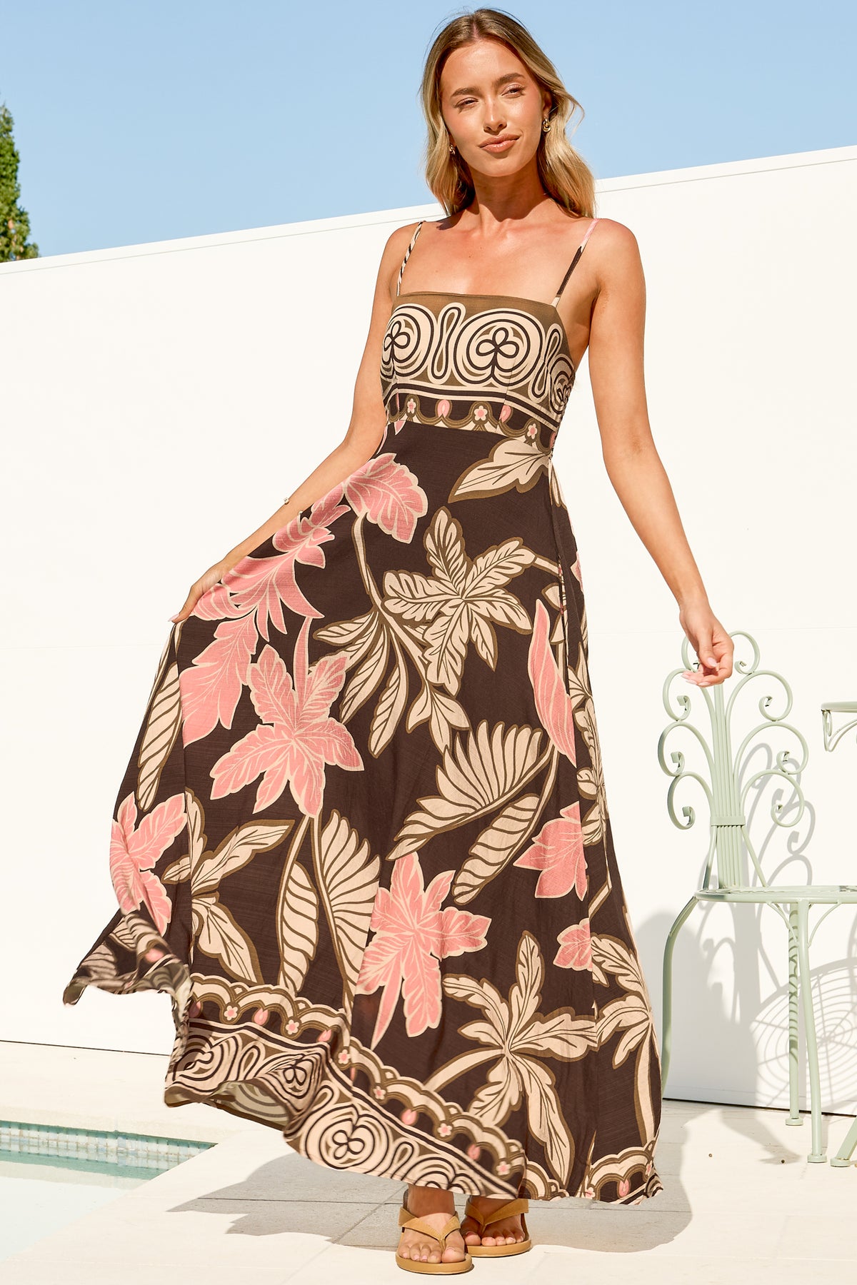 A woman walking and holding out the flowing skirt of the Mattina Maxi Dress in Ginevra Print, a spaghetti strap sun dress in a brown and pink tropical leaf print with a tan swirl border, against a white wall