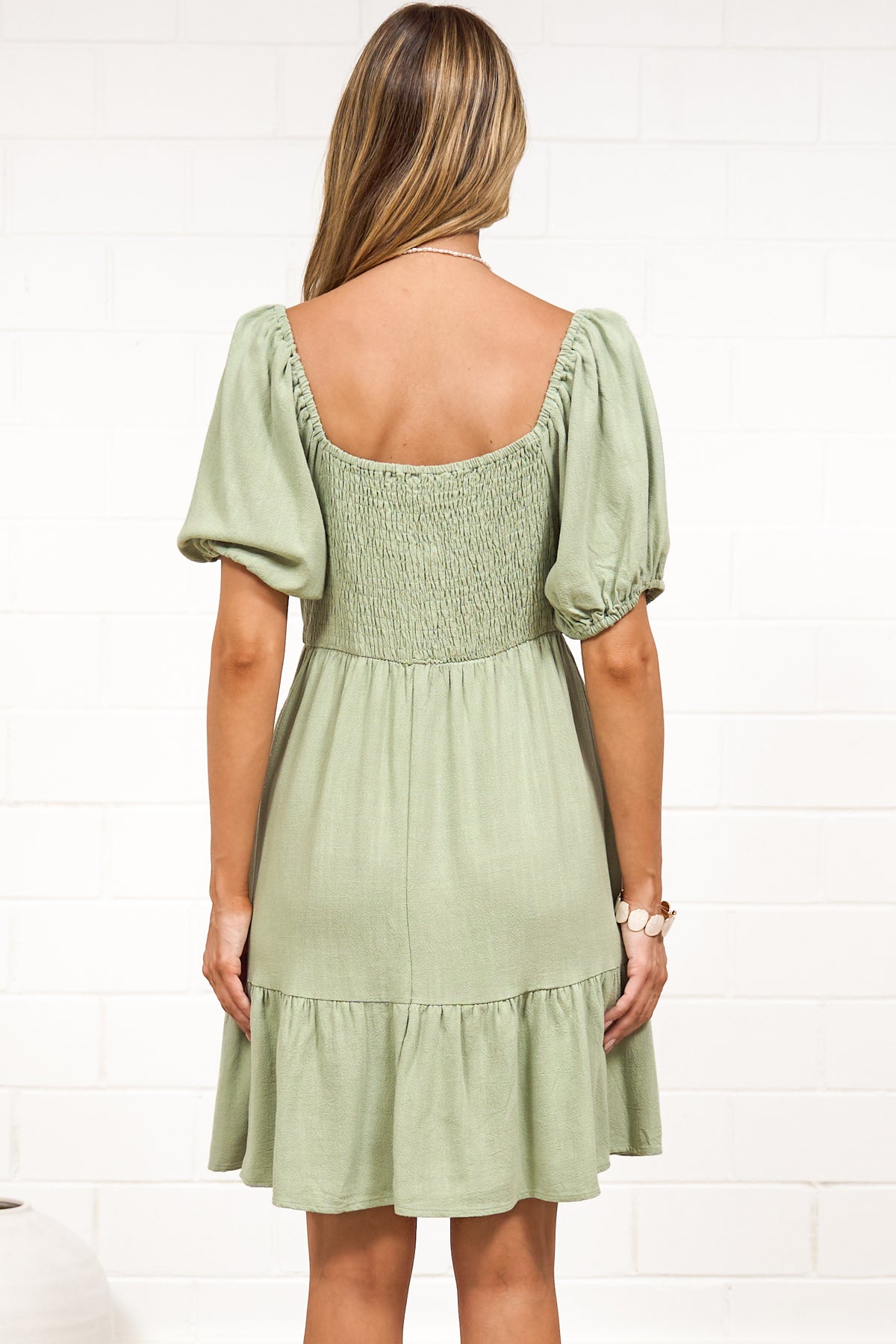 A woman wearing a sage mini dress with a sweetheart neckline, tie-front detail, puff sleeves, and tiered skirt, standing against a white painted brick wall.
