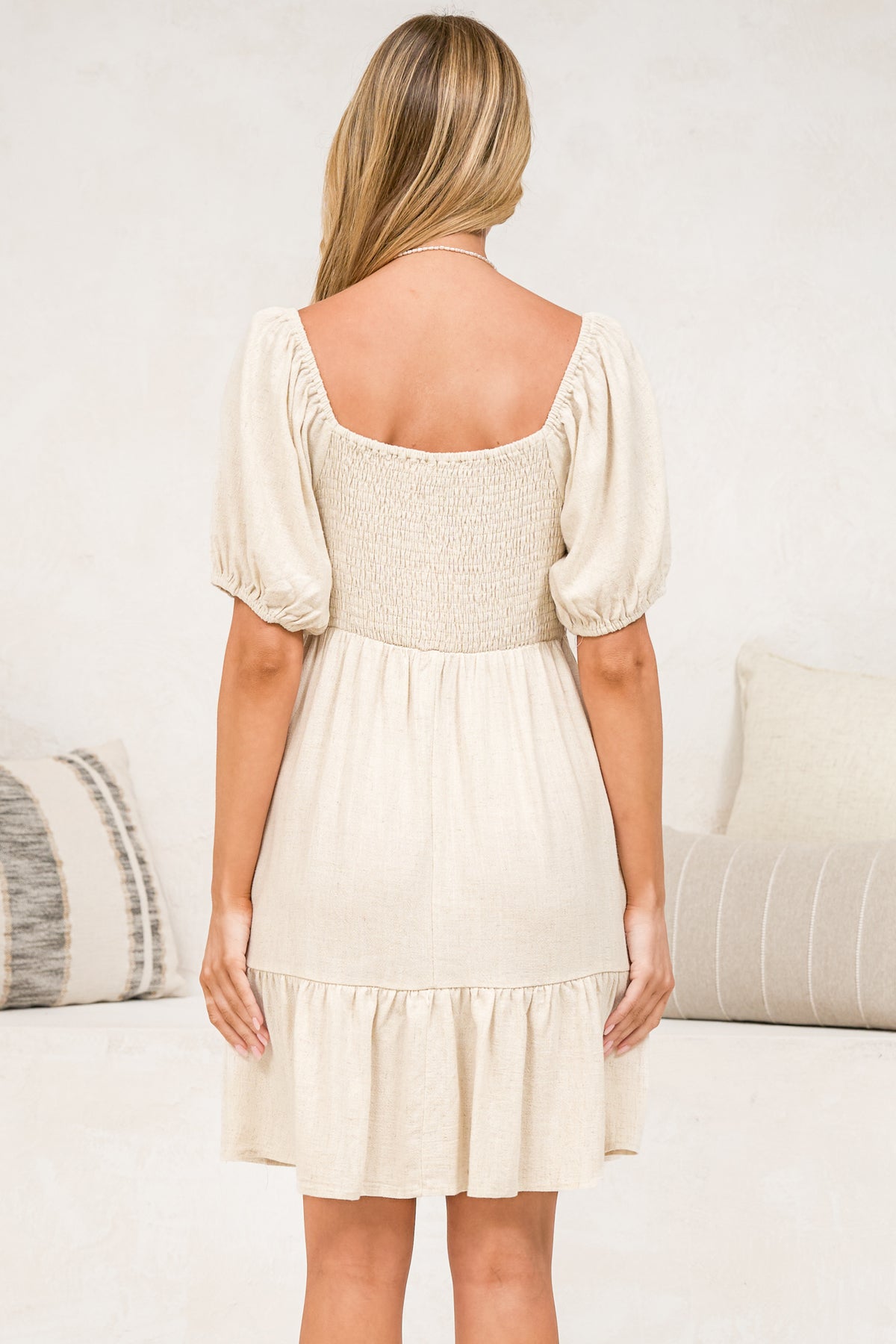 A woman modeling an off-the-shoulder cream-colored mini dress with puff sleeves, a tie-front detail, and a ruffled hem, set against a light-colored indoor background.