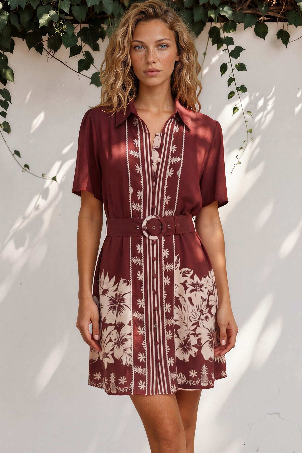 Woman wearing a patterned dress standing against a white wall with greenery.