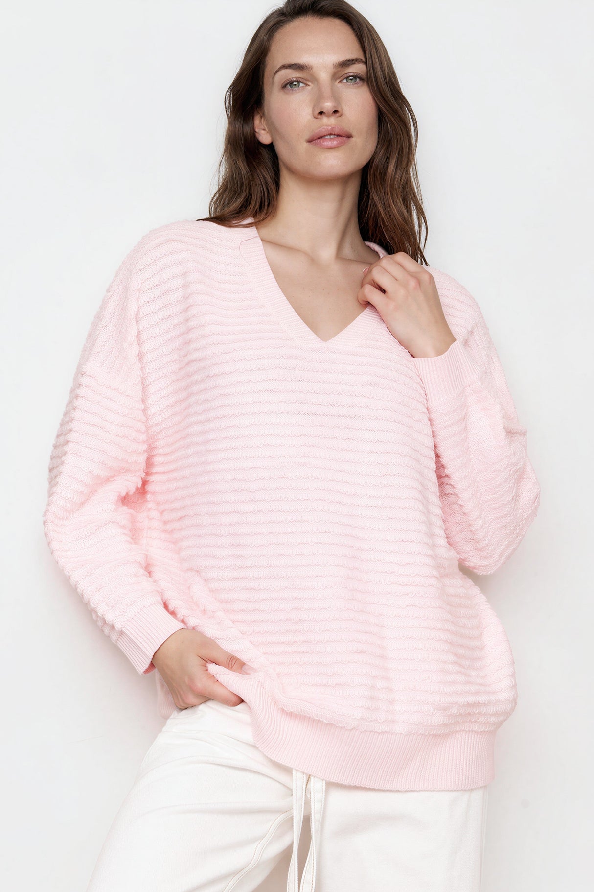Woman wearing a pink ribbed sweater and white pants against a neutral background