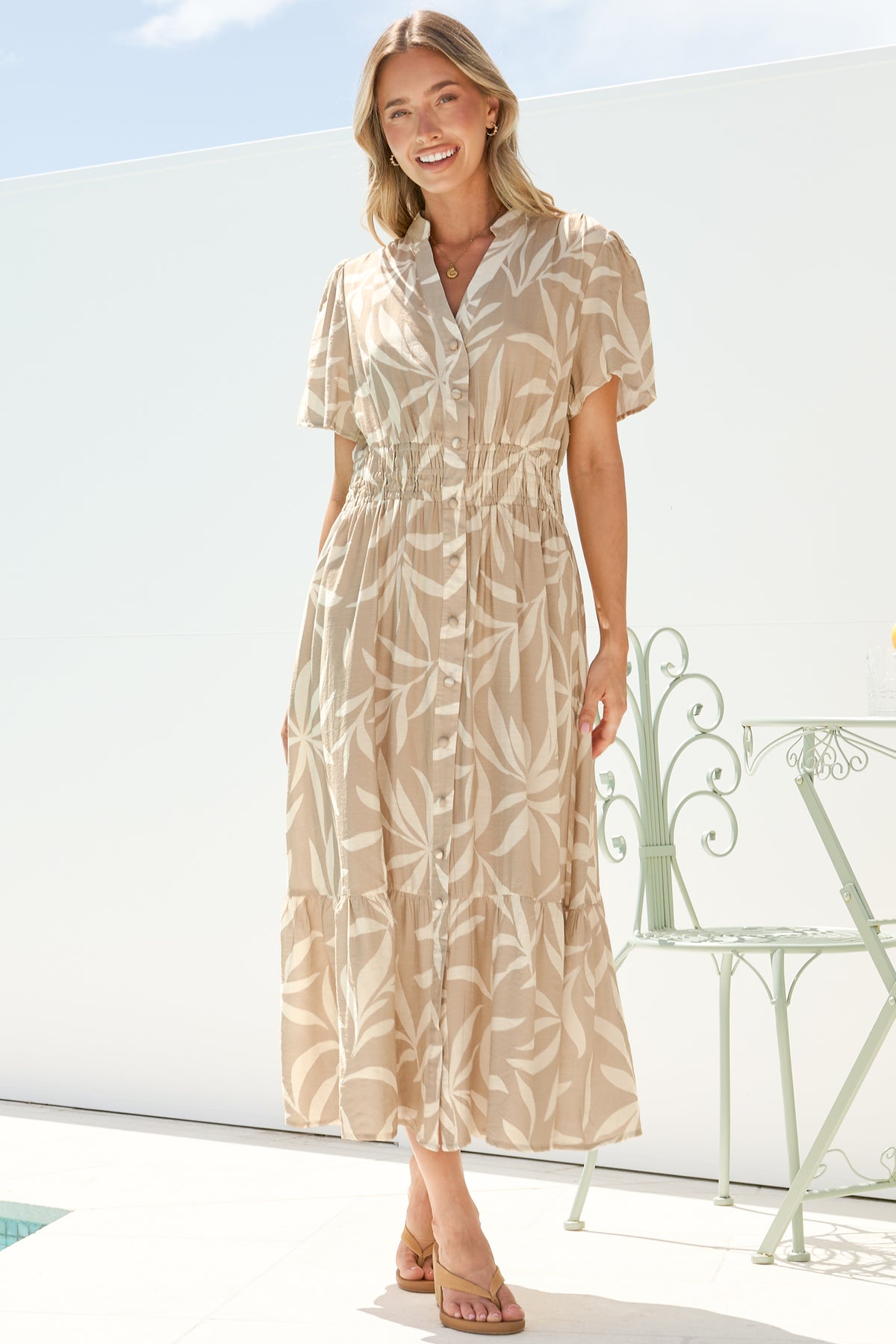 A smiling woman mid-step in the Paloma Midi Dress in Gretchen Print, a flowy beige leaf-print button-down midi with a split mandarin collar and cap sleeves, on a bright white terrace with a garden chair