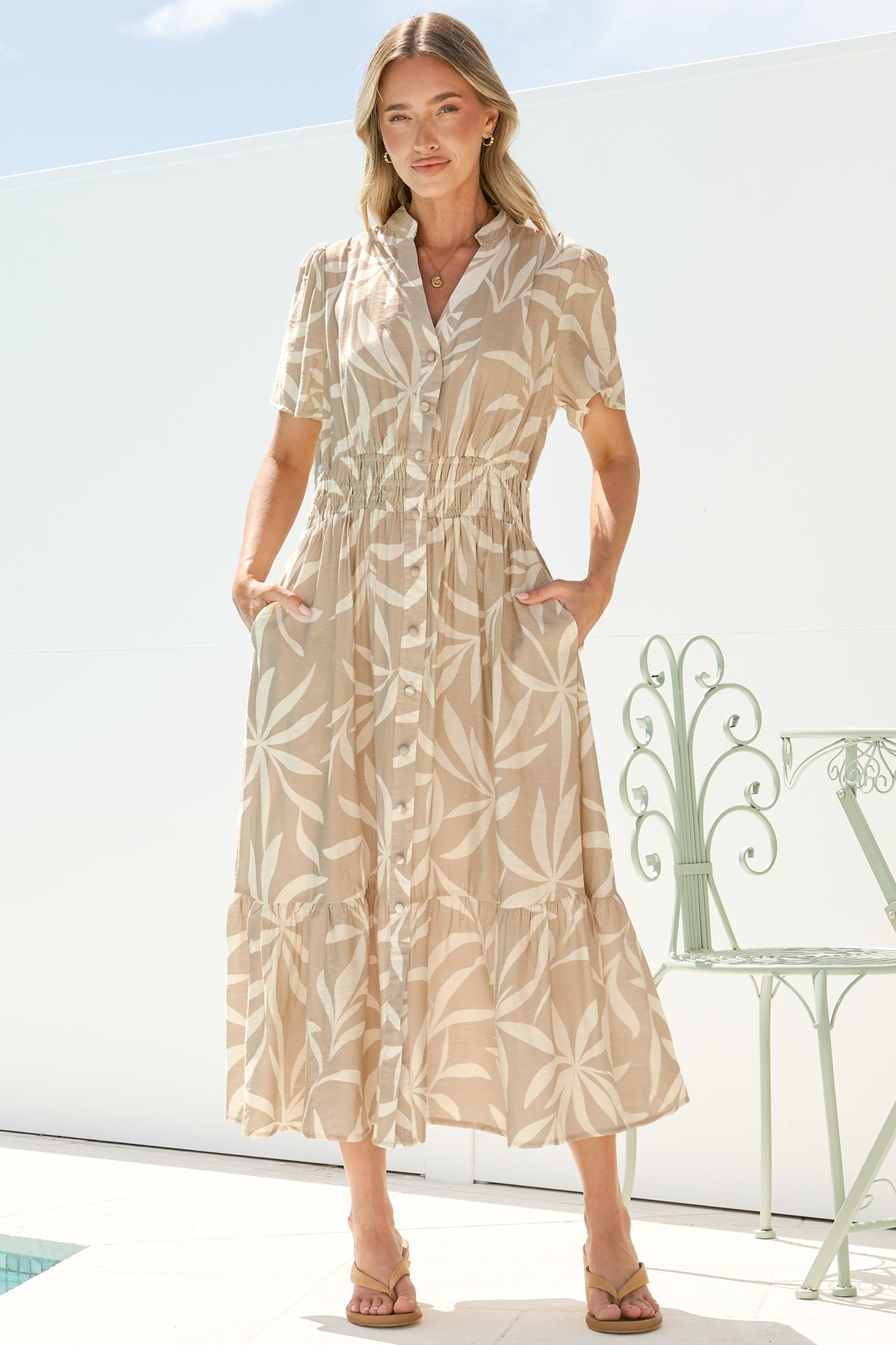 A woman with her hands in the in-seam pockets of the Paloma Midi Dress in Gretchen Print, a beige and cream leaf-print A-line midi with a button front and cap sleeves, on a sunny white terrace