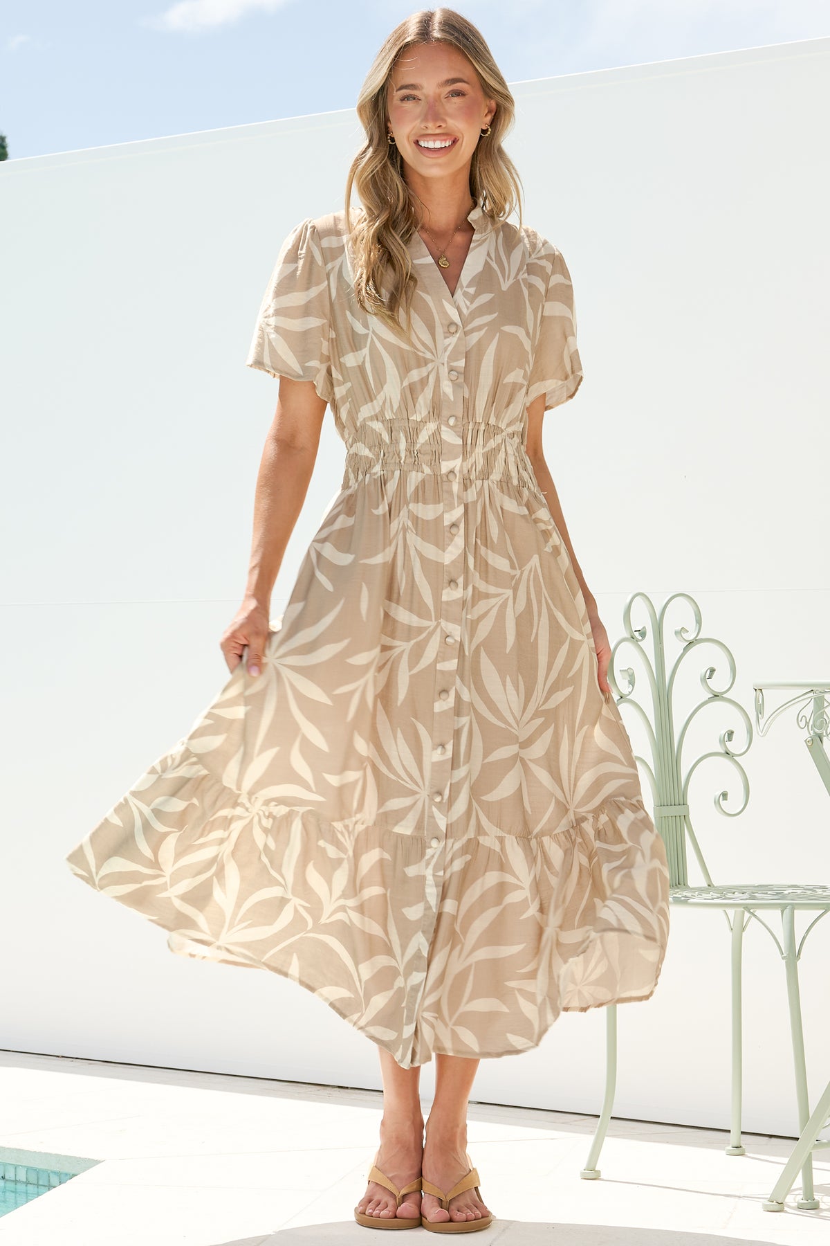 A smiling woman twirling in the Paloma Midi Dress in Gretchen Print, showing the full, swishy A-line skirt of the beige and cream leaf-print button-down midi, on a sunny white terrace with a garden chair