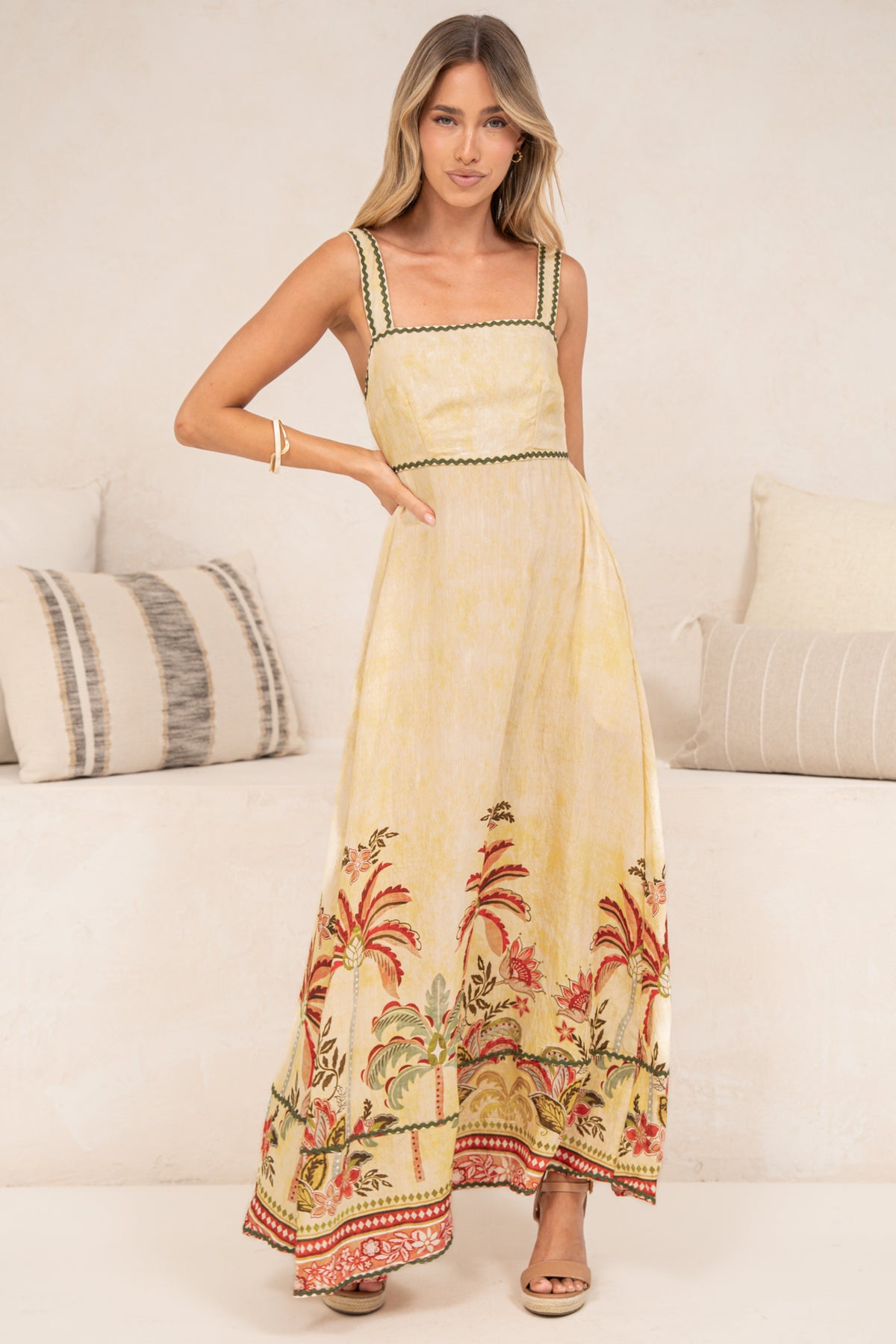 A woman posing with one hand on her hip in the Poppy Maxi Dress in Palmina Print, showing the butter yellow fitted bodice, ric rac trims and flowing maxi skirt with a red and green palm tree border, styled with tan sandals in front of a cream sofa