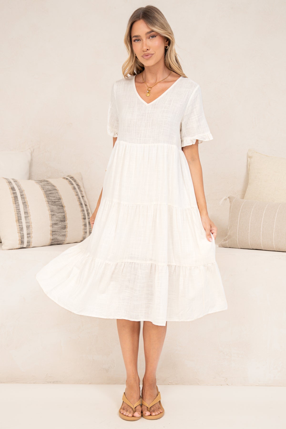 A woman holding out the skirt of the Porcelaine Midi Dress in white to show its flowy tiered midi shape, with a V-neckline and frill-trimmed short sleeves, standing in tan sandals in front of a cream sofa with neutral cushions