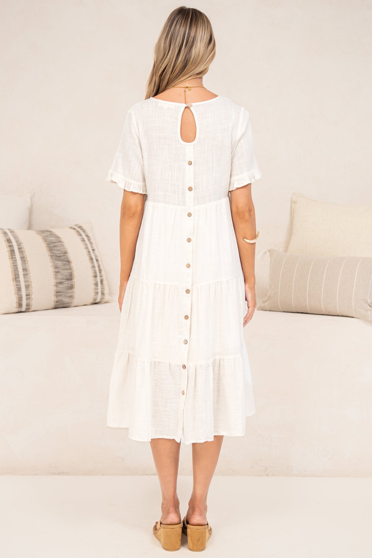 Back view of a woman wearing the Porcelaine Midi Dress in white, showing the keyhole neckline, wooden button closure down the back and tiered midi skirt, styled with tan sandals in front of a cream sofa with neutral cushions