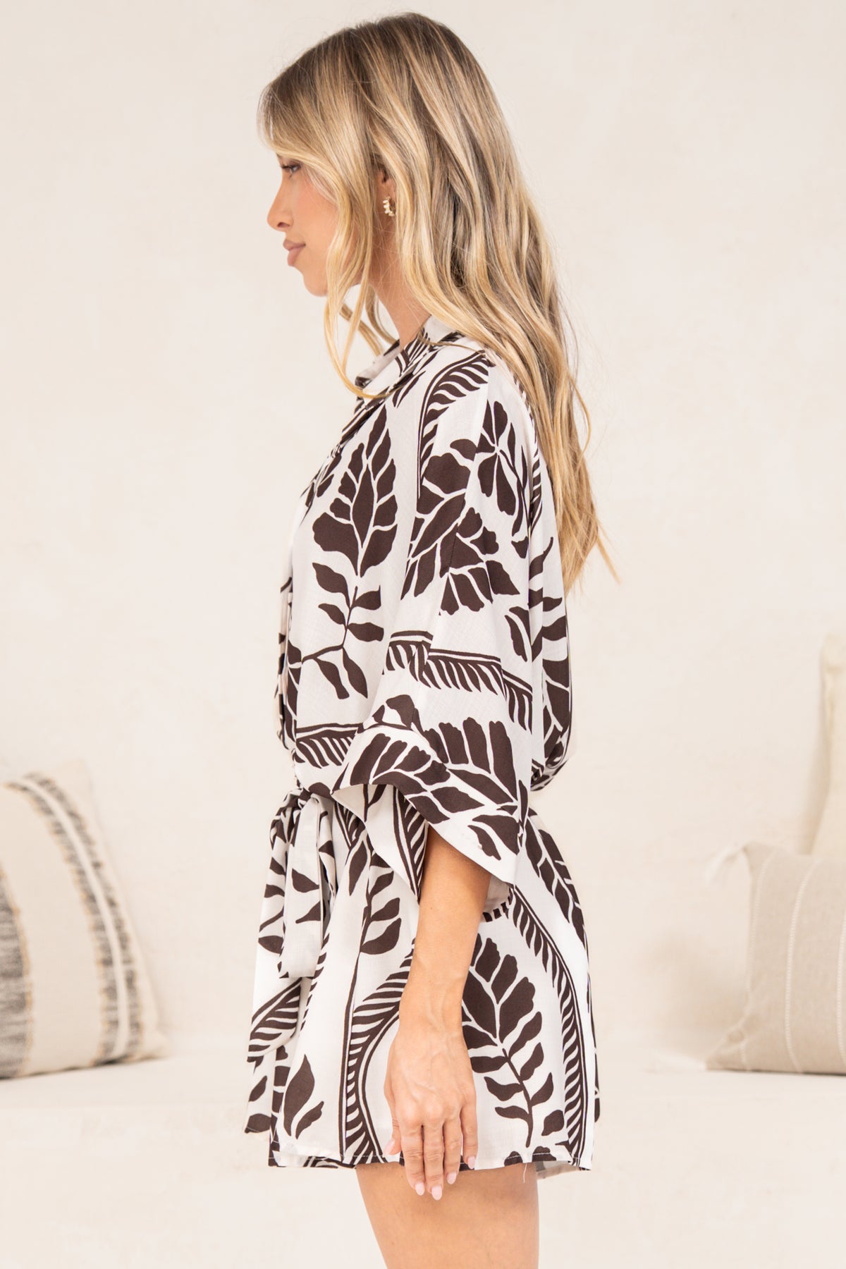 A woman in a tropical-inspired printed playsuit featuring wide sleeve cuffs, an elasticated waist, and relaxed shorts, posed beside a cream couch.