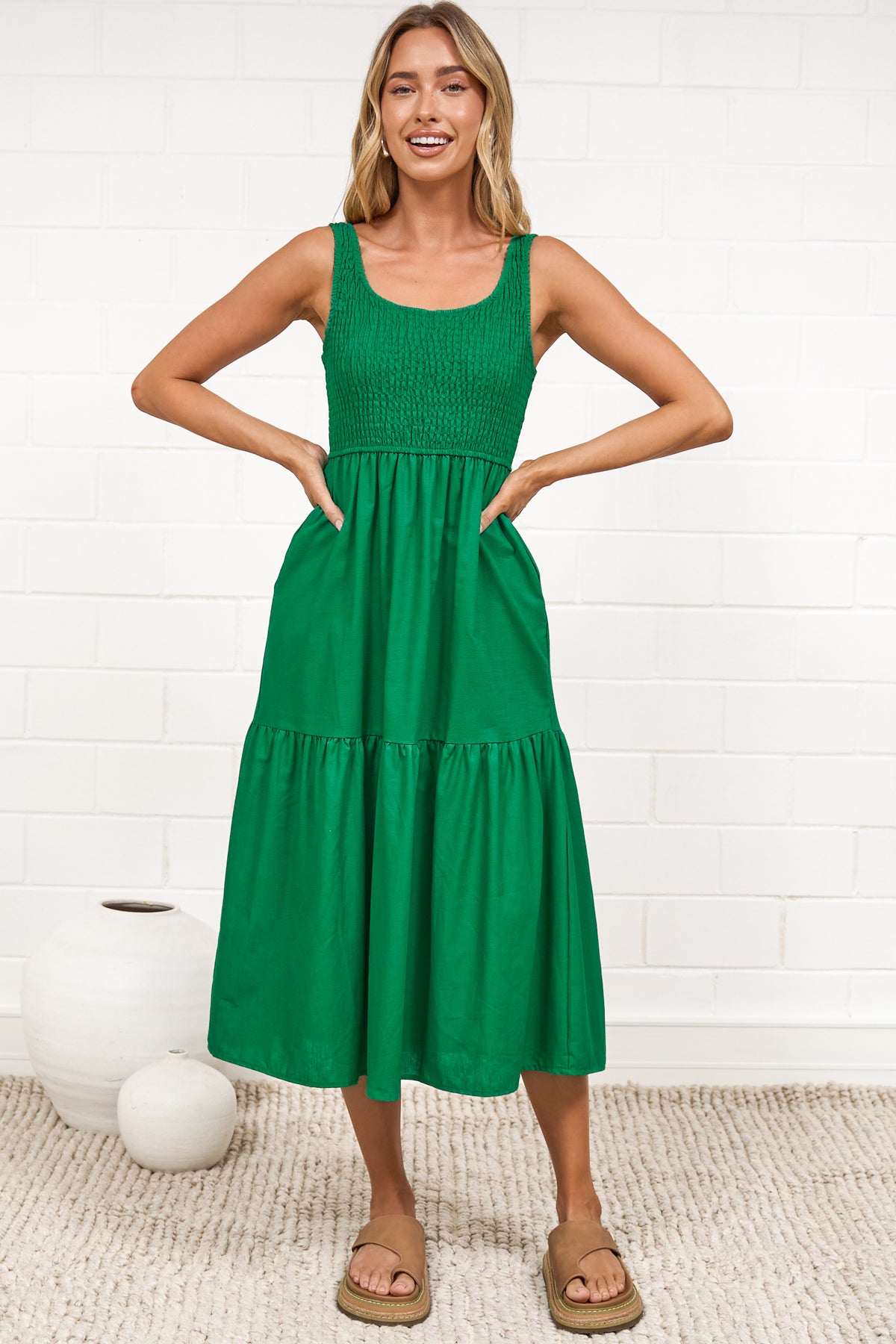 A woman in a flowing green midi dress featuring a shirred bodice and tiered skirt, posed against a white painted brick wall.