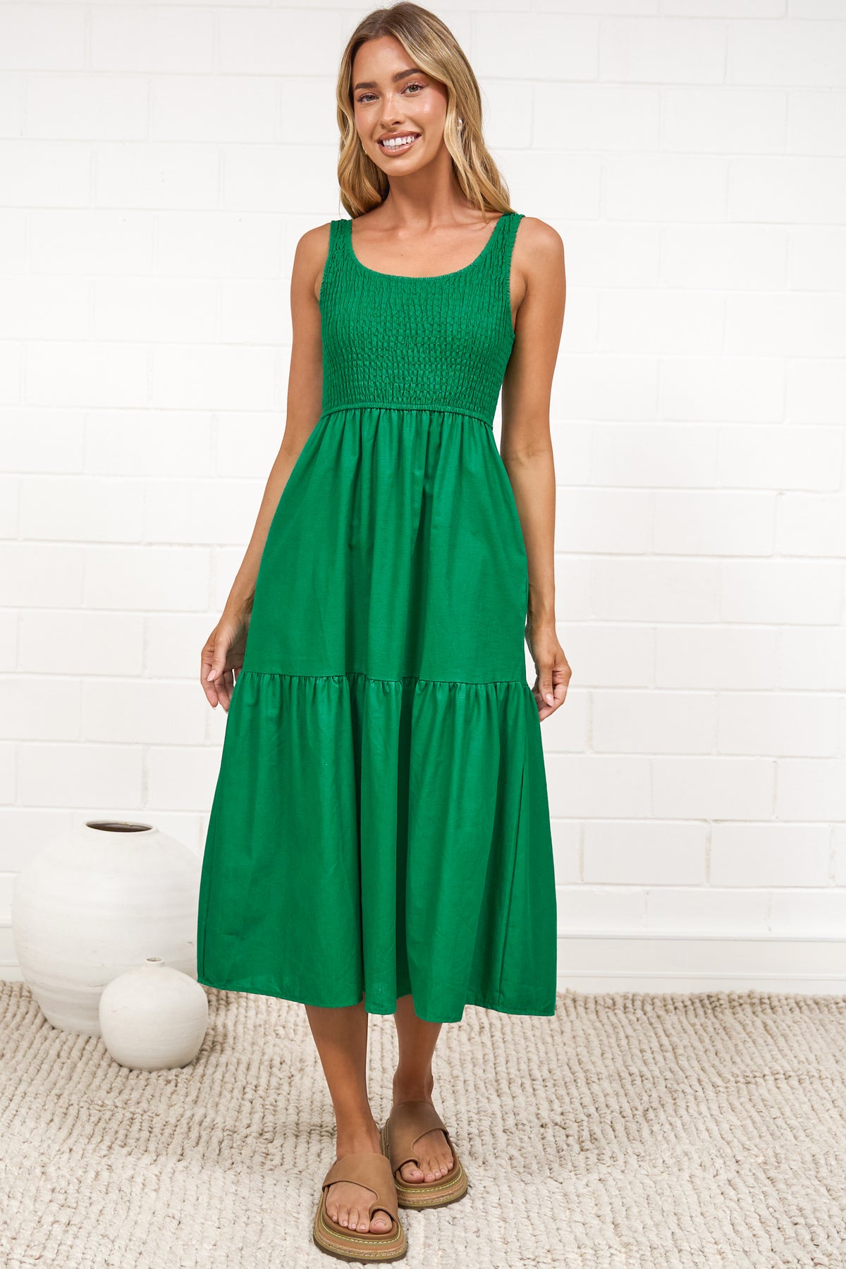 A woman in a flowing green midi dress featuring a shirred bodice and tiered skirt, posed against a white painted brick wall.