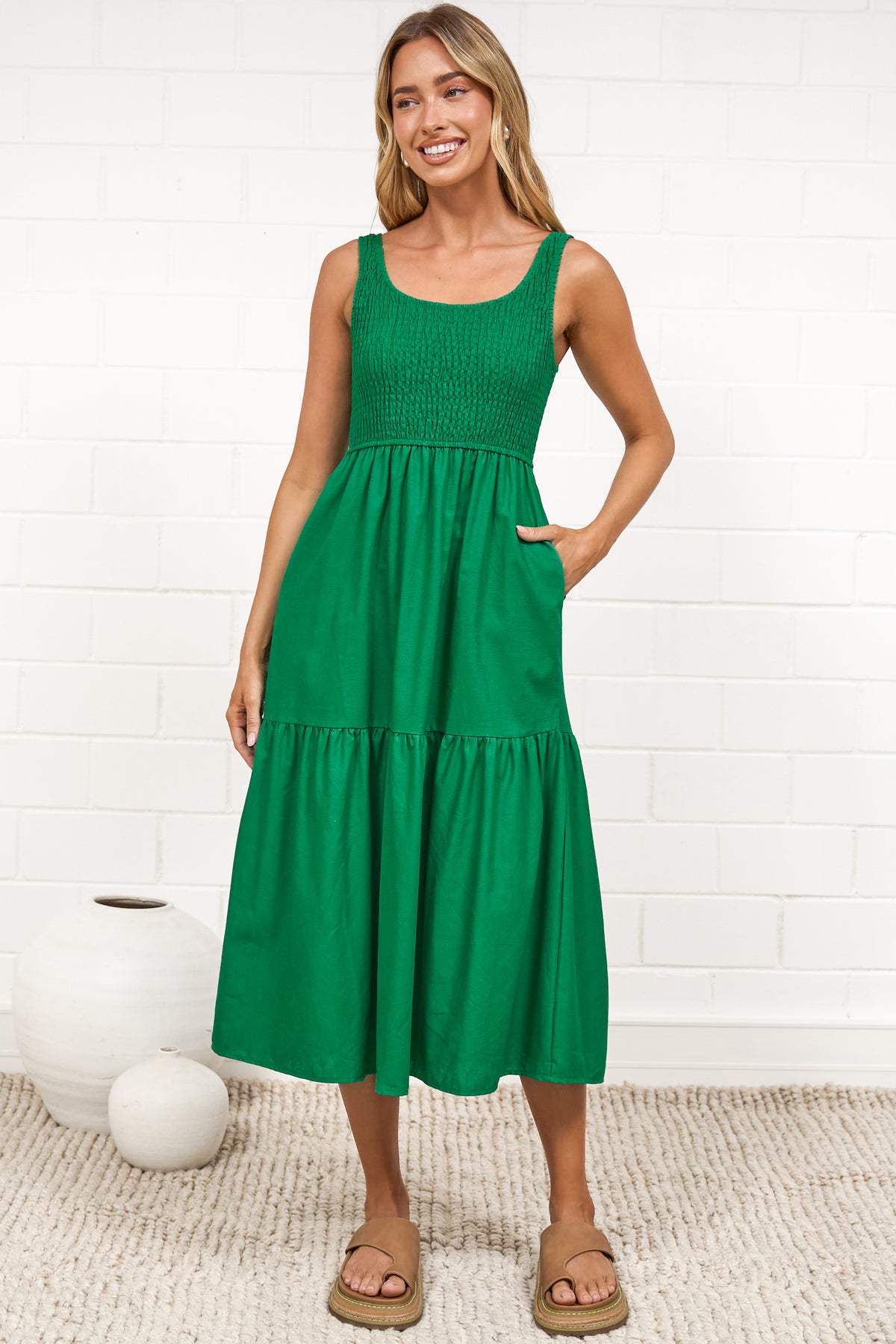 A woman in a flowing green midi dress featuring a shirred bodice and tiered skirt, posed against a white painted brick wall.