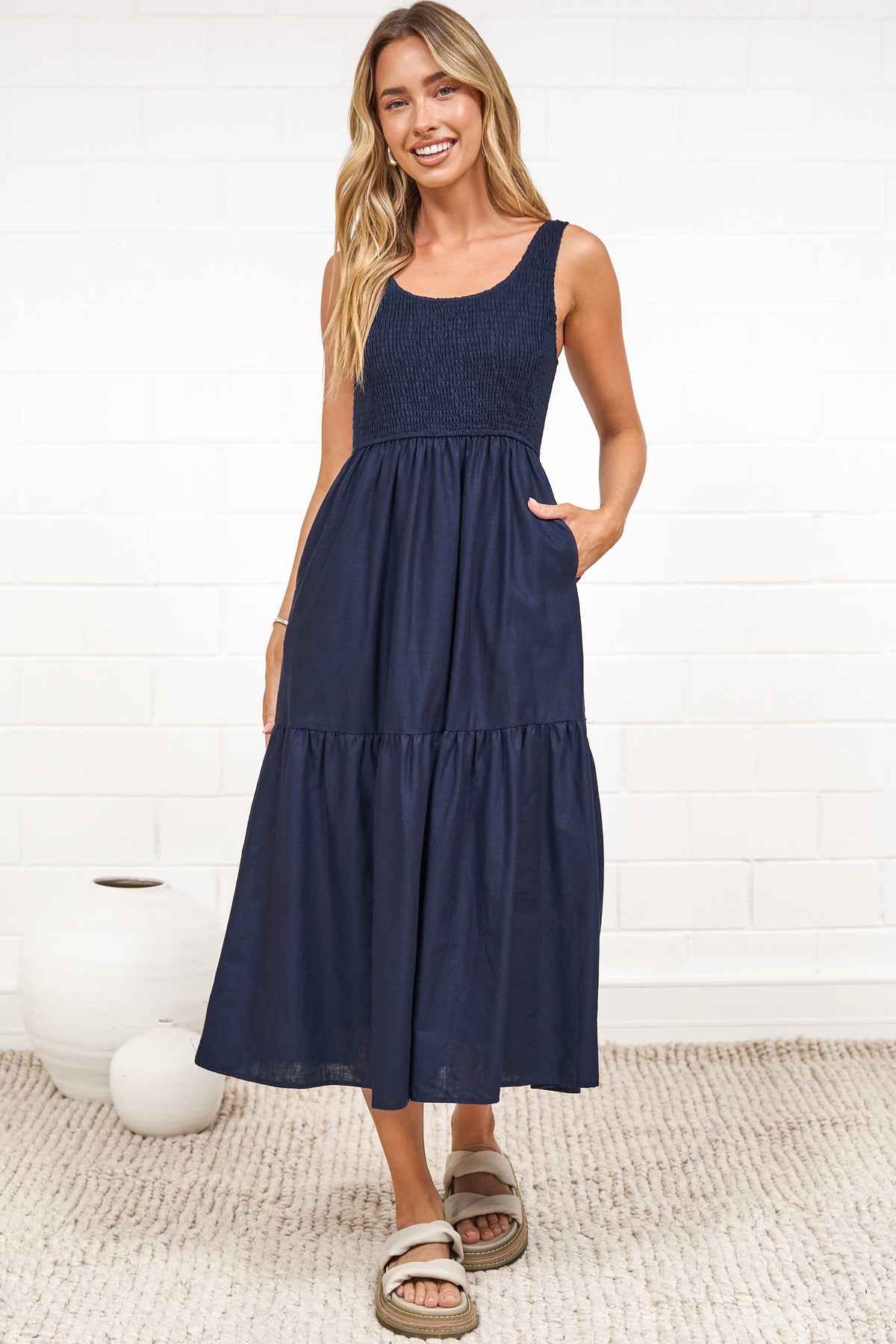 A woman in a rust midi dress with thick straps, a shirred bodice, and tiered skirt, posed against a white painted brick wall.