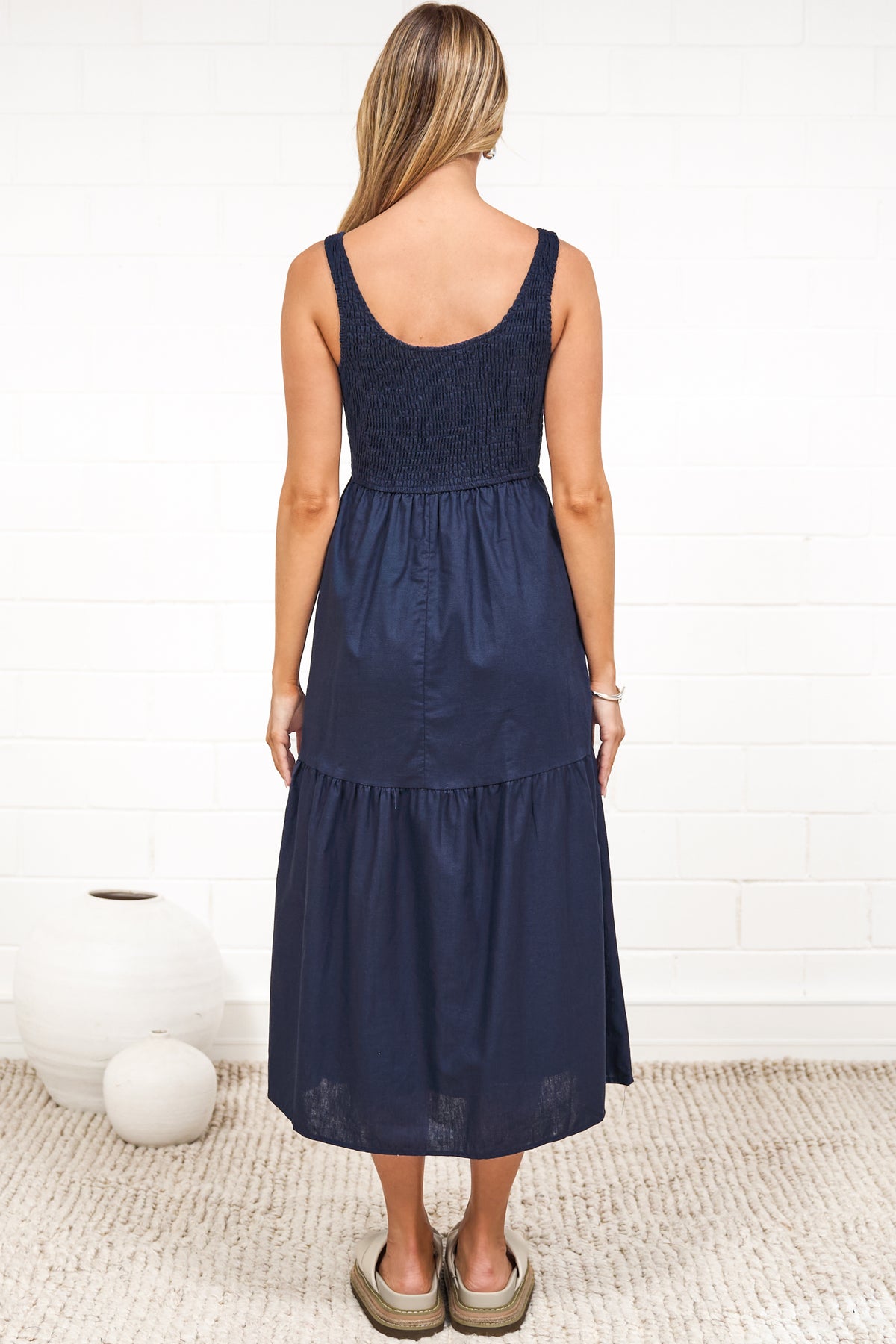A woman in a rust midi dress with thick straps, a shirred bodice, and tiered skirt, posed against a white painted brick wall.