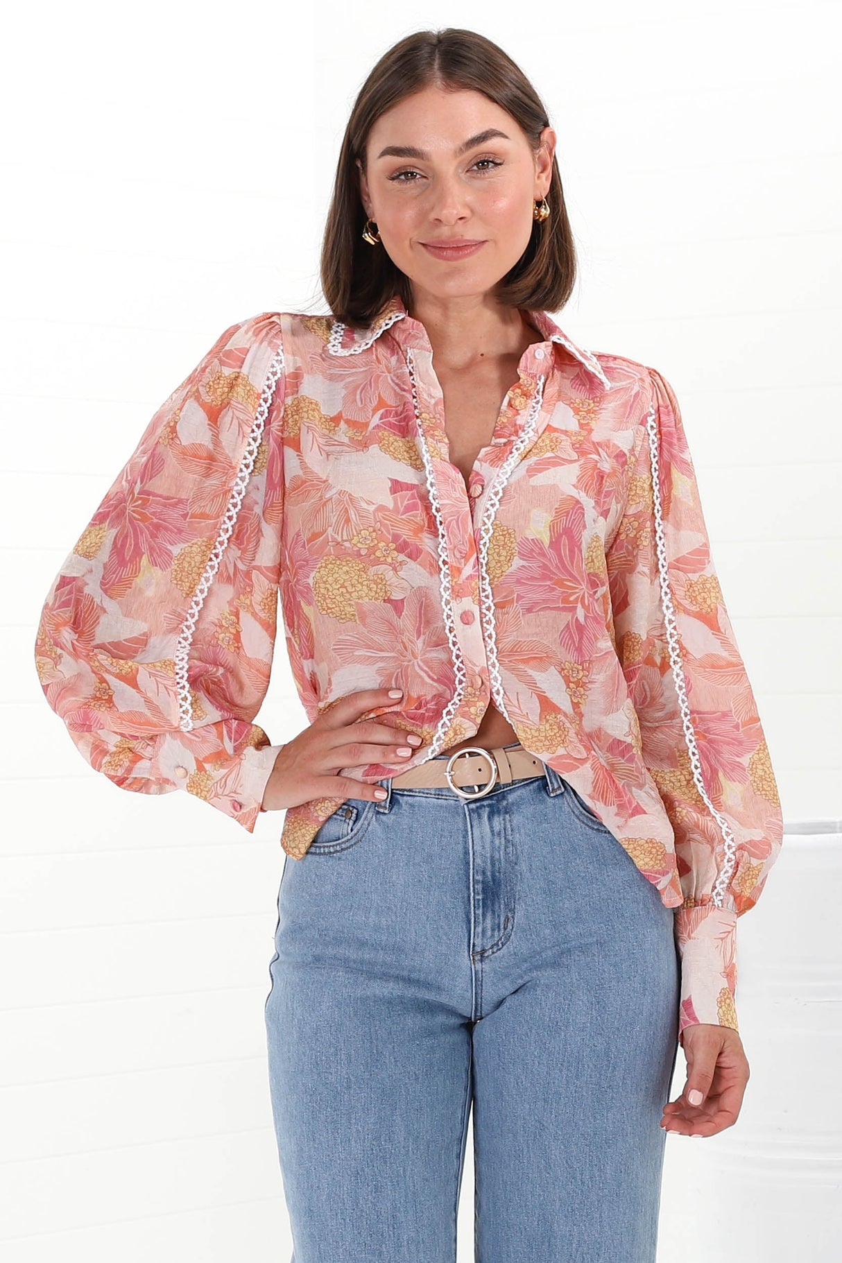 A woman wearing a pink and orange floral button-down blouse with contrast trim lace detailing, with long sleeves and a scoop hemline, paired with blue jeans.