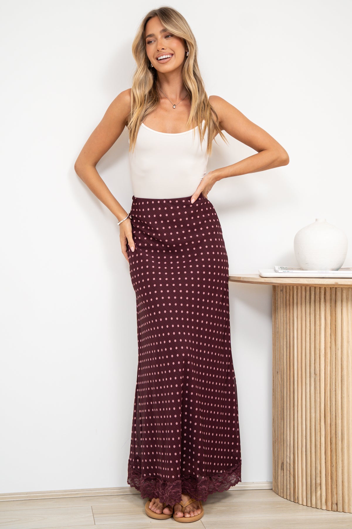 A woman in a wine maxi skirt featuring a delicate polka dot print, sleek bias-cut silhouette, and lace-trimmed hem, posed against an off-white painted wall.