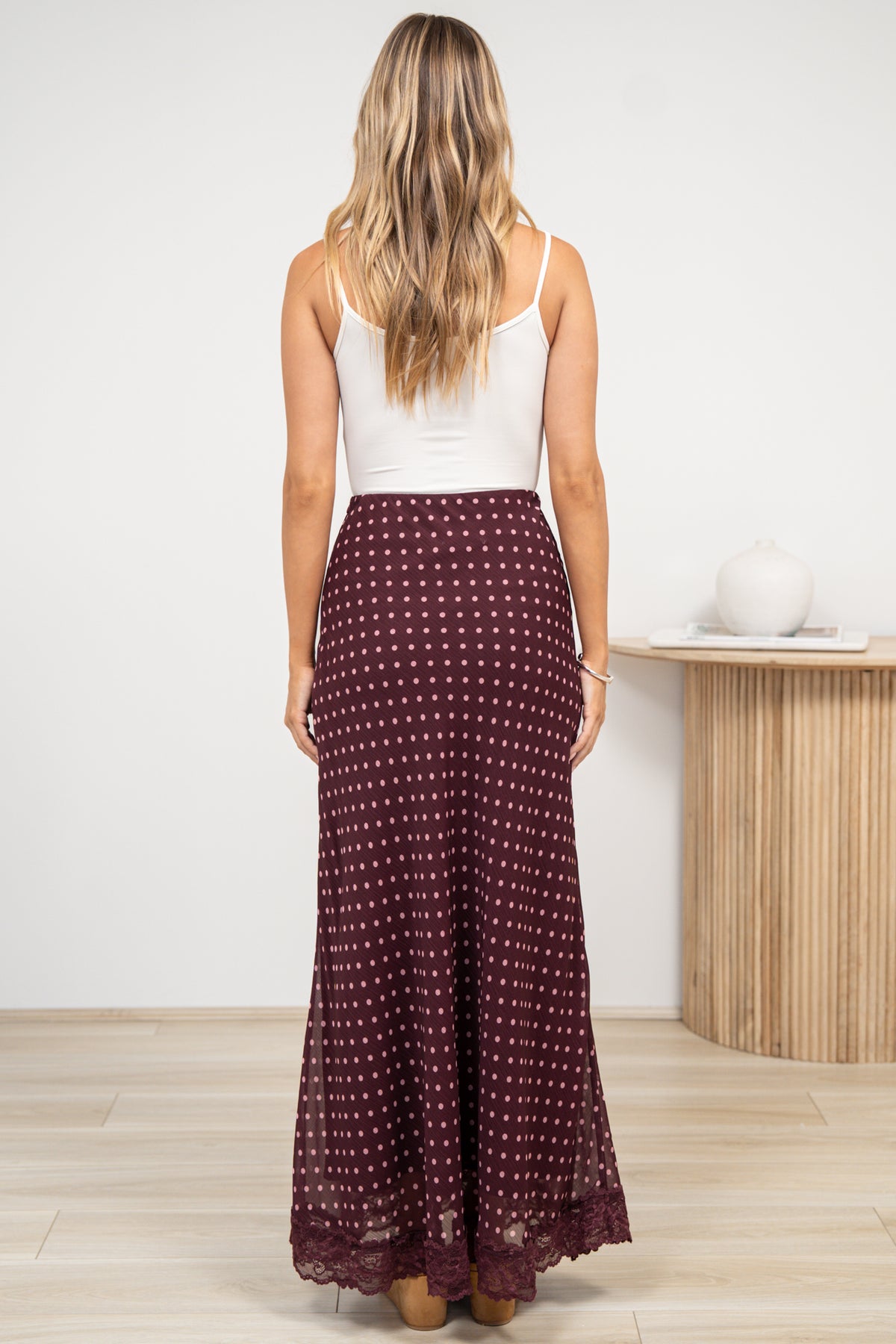 A woman in a wine maxi skirt featuring a delicate polka dot print, sleek bias-cut silhouette, and lace-trimmed hem, posed against an off-white painted wall.