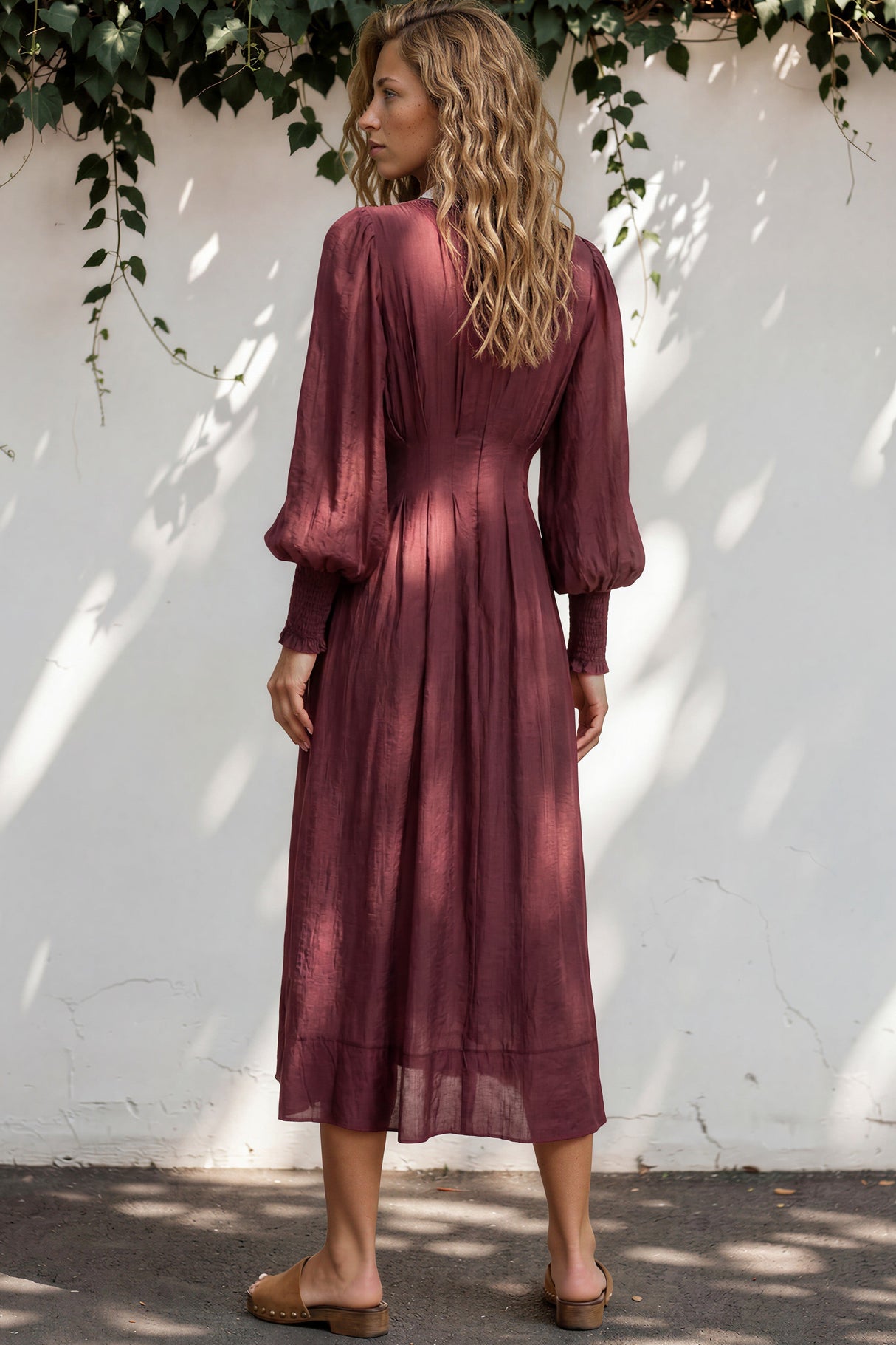 Woman wearing a burgundy dress standing against a white wall with greenery.
