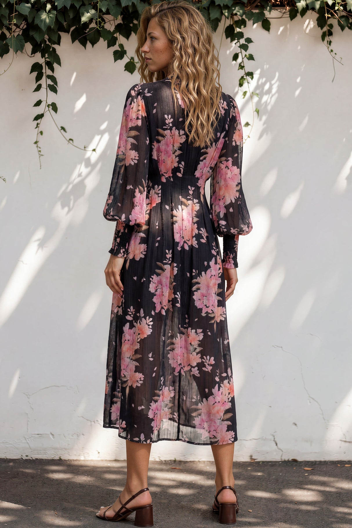 Woman wearing a floral dress standing against a white wall with greenery.