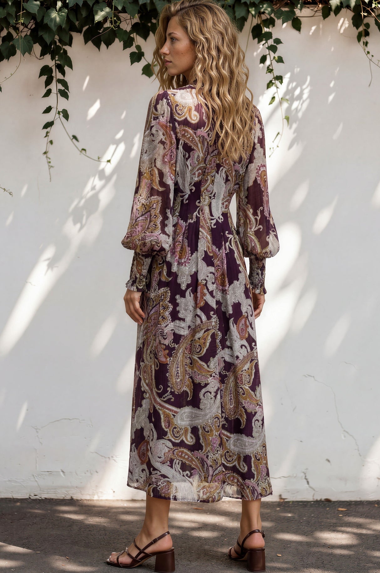 Woman wearing a patterned dress standing against a white wall with greenery.