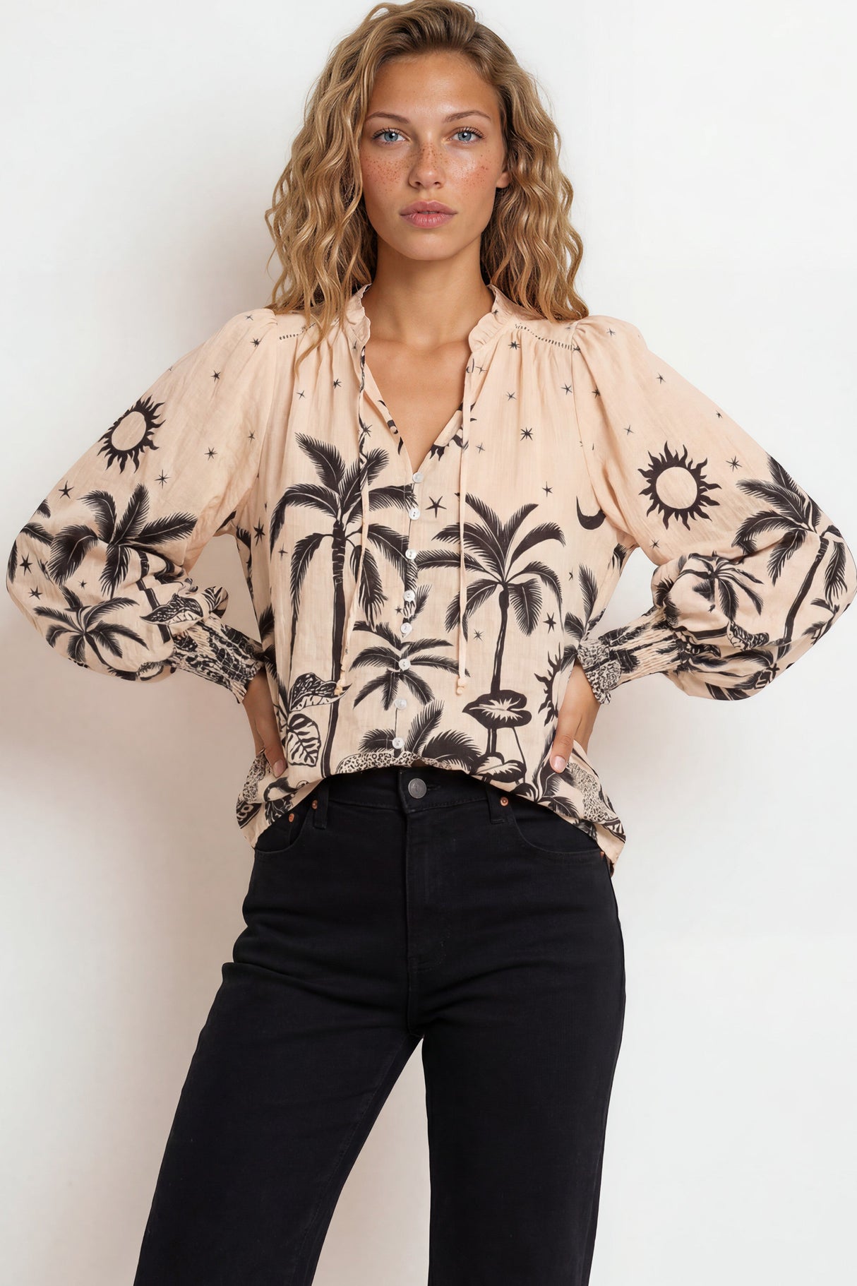 Woman wearing a beige blouse with black palm tree pattern and black pants against a white background