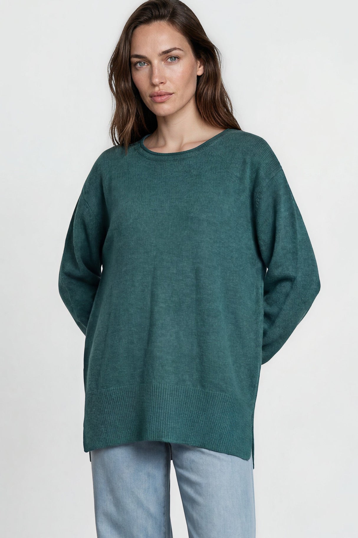 Woman wearing a green sweater and light blue jeans standing against a white wall.