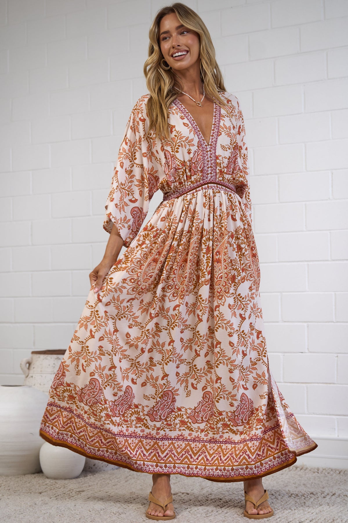A woman modeling a bohemian-style maxi dress with a warm orange, pink, and beige floral pattern, standing in a bright room with a white brick wall and a textured rug.