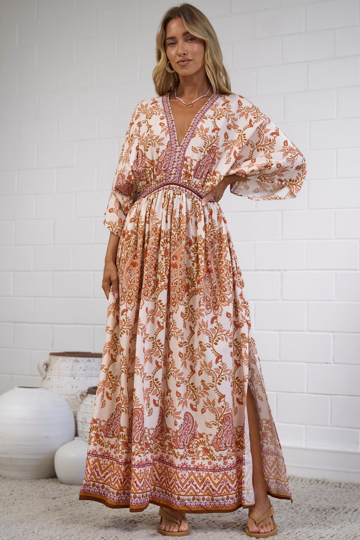 A woman modeling a bohemian-style maxi dress with a warm-toned floral and geometric pattern in orange, rust, and beige, standing in a room with a white brick wall and neutral decor.
