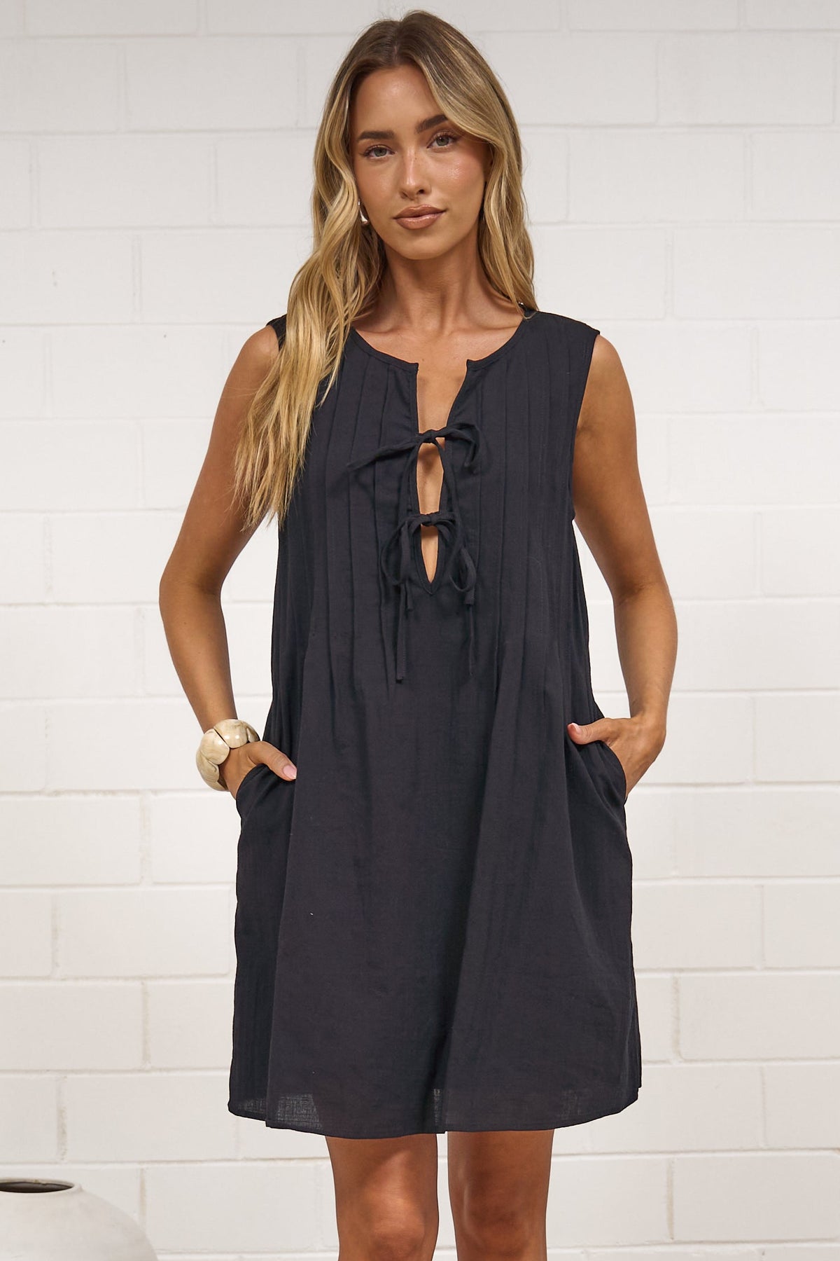 A woman modeling a sleeveless black dress with a keyhole cutout and side pockets, standing on a textured beige rug in front of a white brick wall.