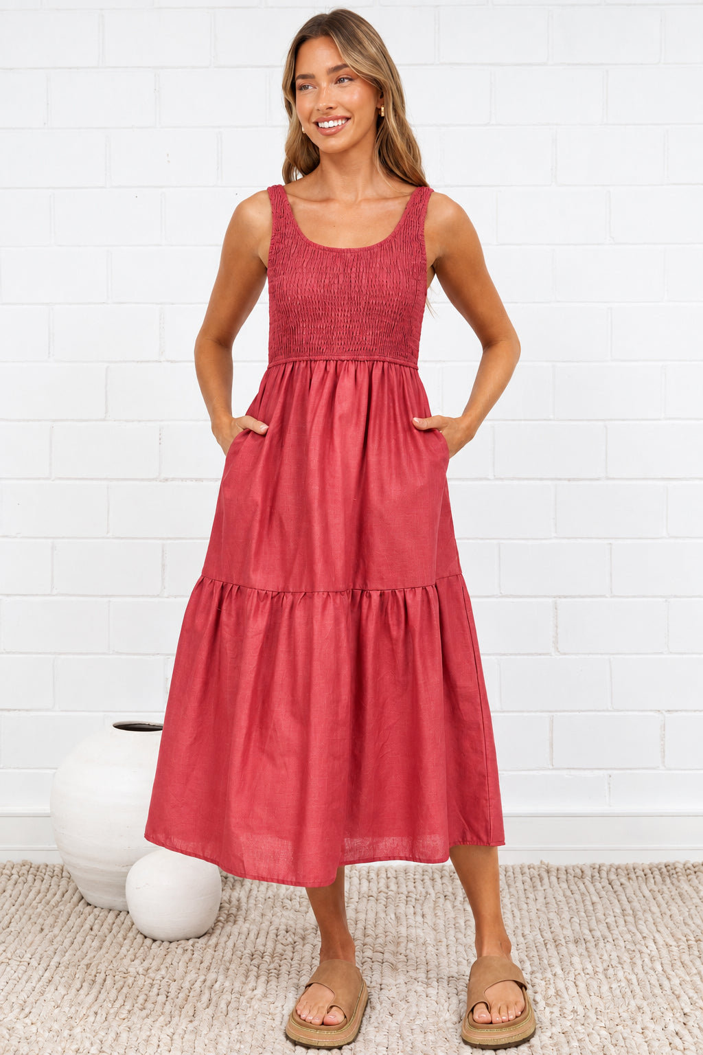 A woman stands in a bright room wearing a sleeveless, tiered midi dress in a dusty rose or terracotta pink color. The dress has a square neckline and a gathered waist with a flared skirt.