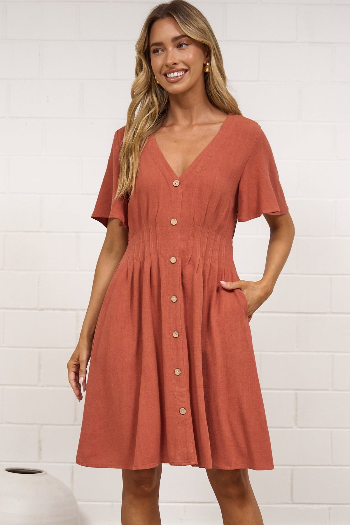 A woman modeling a short-sleeve terracotta dress with a button-down front, standing on a woven rug against a white brick wall.