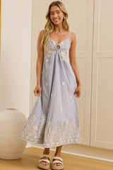 A woman stands in a bright room wearing a light blue sleeveless maxi dress with white floral embroidery and a gathered bow at the neckline. She is posed in front of a large floor mirror that reflects her side profile, with cream paneled walls, wooden flooring, and a white vase in the background. 
