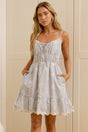 A woman wearing a light blue and white tiered ruffle dress stands in a bright room with cream-colored cabinets, with a large mirror on the left reflecting her side profile.
