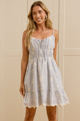 A woman stands in a bright room wearing a light blue sleeveless tiered dress with white floral embroidery. She is standing in front of cream-colored wardrobe doors, with a large mirror on the left reflecting her side profile. 