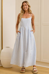 A woman stands in a bright room wearing a light blue and white gingham maxi dress with side pockets, with a large mirror on the left reflecting her profile. The room has cream-colored paneled walls and doors, light wood floors, and two white ceramic vases.