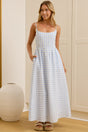 A woman modeling a light blue and white gingham maxi dress with spaghetti straps and side pockets, standing in a bright room with cream-colored cabinets and a large mirror showing her reflection.