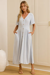 A woman modeling a light blue midi dress with short sleeves, a V-neckline, and front buttons, standing in a room with cream-colored cabinets and a mirror showing her side profile.