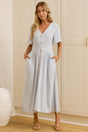 A woman modeling a light blue midi dress with short sleeves, a V-neckline, and front buttons, standing in a room with cream-colored cabinets and a mirror showing her side profile.