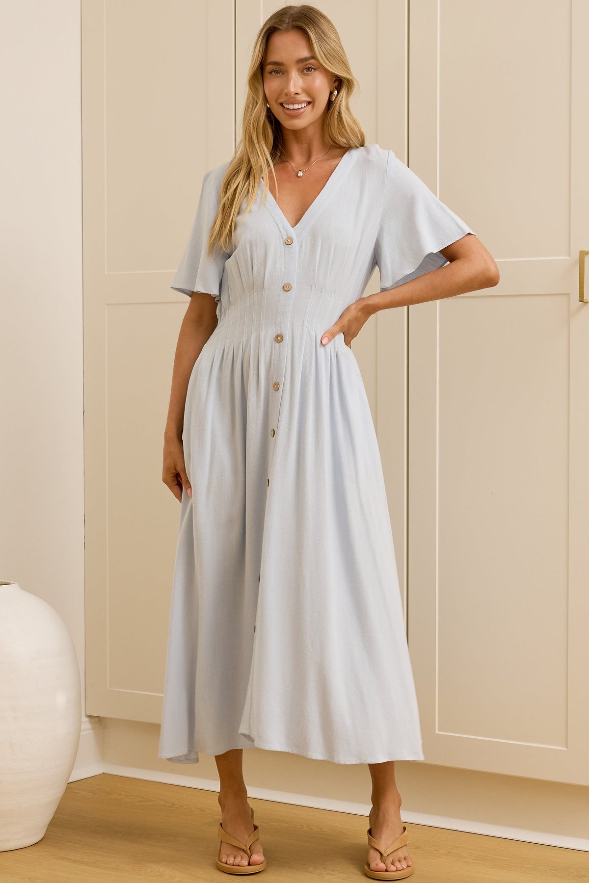 A woman modeling a light blue midi dress with flutter sleeves and a button-front bodice, standing in a bright room with cream-colored wardrobes and a wooden floor. Her reflection is visible in a mirror on the left.