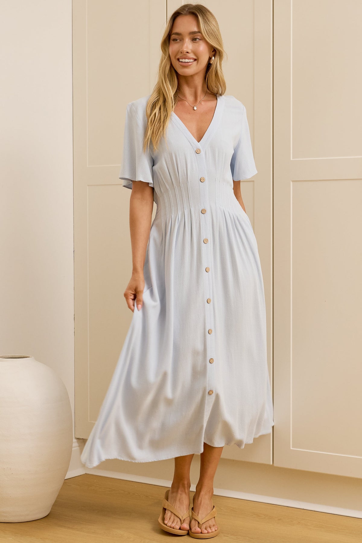 A woman wearing a light blue midi dress with short sleeves and a button-down front stands in a bright room with cream-colored wardrobes and a wooden floor. A large mirror on the left reflects her side profile.