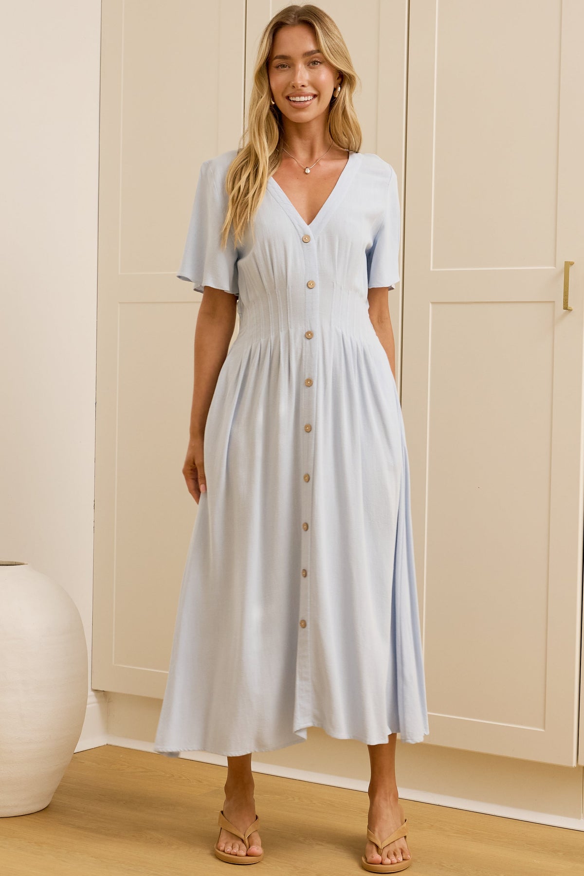 A woman modeling a light blue midi dress with short sleeves and a button-down front, standing in a bright room with cream-colored cabinets and a mirror showing her side profile.