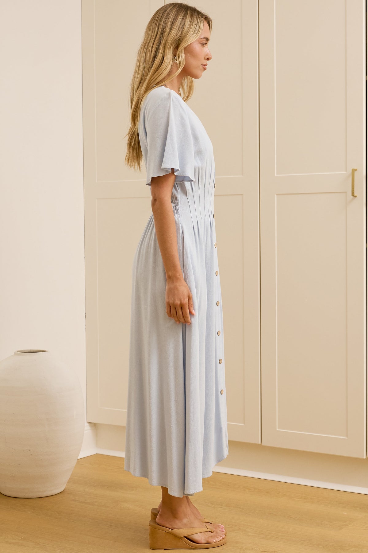 A woman stands in profile wearing a light blue midi dress with short sleeves and a button-front detail, set against a background of cream-colored wardrobe doors and a large mirror reflecting her back.